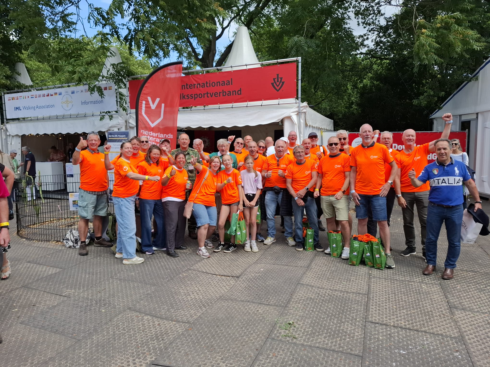 Groepsfoto van ongeveer dertig deelnemers in feloranje shirts voor een evenementenpaviljoen tijdens de Nijmeegse Vierdaagse. De groep staat buiten onder bomen en poseert voor een stand van het NLVi