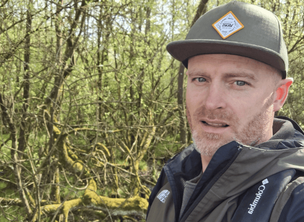 Een persoon met een groene pet en outdoorjas staat in een bosrijke omgeving met mosbegroeide takken en jonge lentebegroeiing op de achtergrond. De persoon kijkt recht in de camera terwijl hij zich in de natuur bevindt tijdens een wandeling of verkenningstocht.