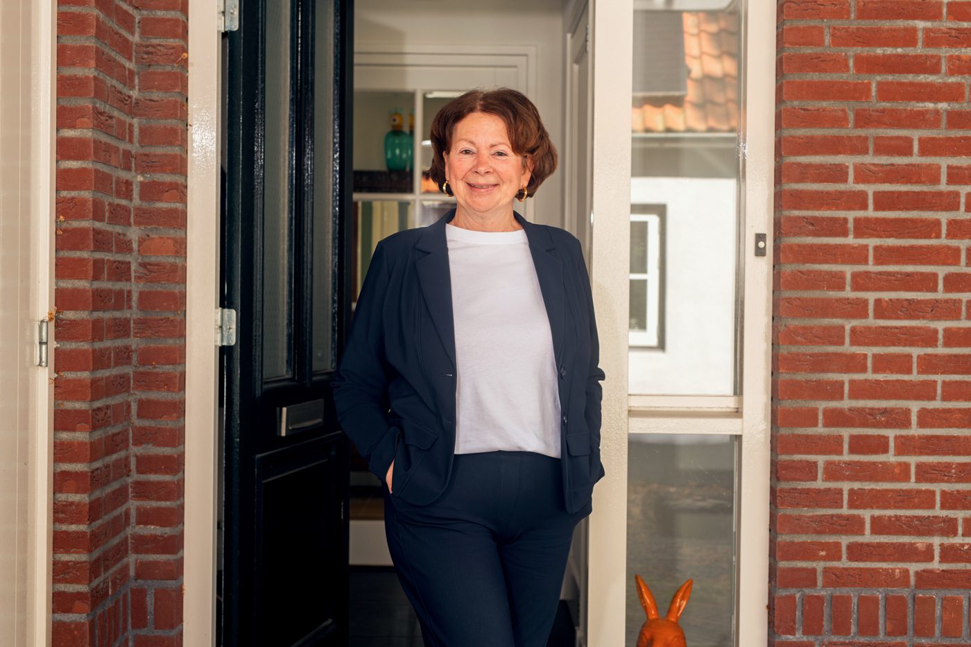 Persoon staat in de open voordeur van een bakstenen woning en kijkt in de richting van de camera. De persoon draagt een donkerblauw pak met een licht shirt en heeft de handen in de zakken. Op de achtergrond is een deel van de hal zichtbaar. Rechts onderin staat een oranje decoratief beeldje van een haas. De foto is gemaakt bij daglicht en toont de entree van de woning als achtergrond.