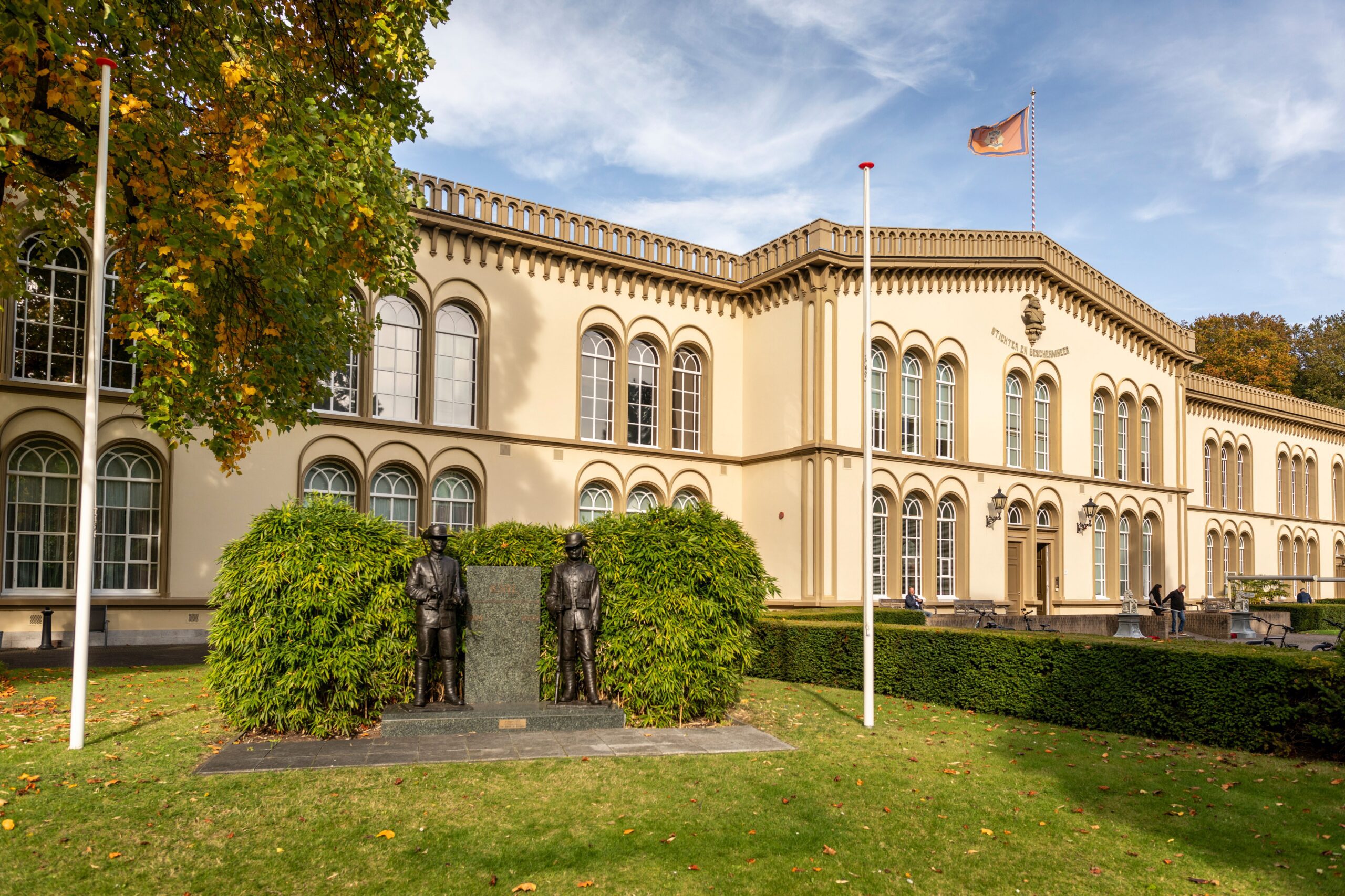 Vooraanzicht van het Koninklijk Tehuis voor Oud-Militairen en Museum Bronbeek, met een monument op het gazon, bomen en een vlag voor het gebouw.