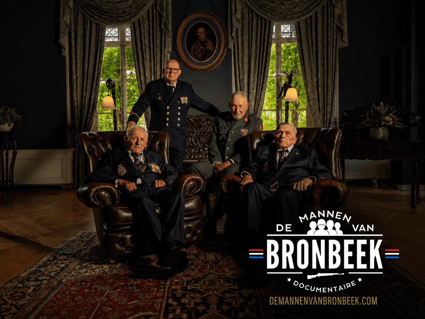 Vier oudere veteranen poseren in uniform in een klassieke, donker ingerichte kamer. Twee mannen zitten in leren fauteuils en twee staan achter hen. Rechtsonder staat het logo van de documentaire ‘De Mannen van Bronbeek’.