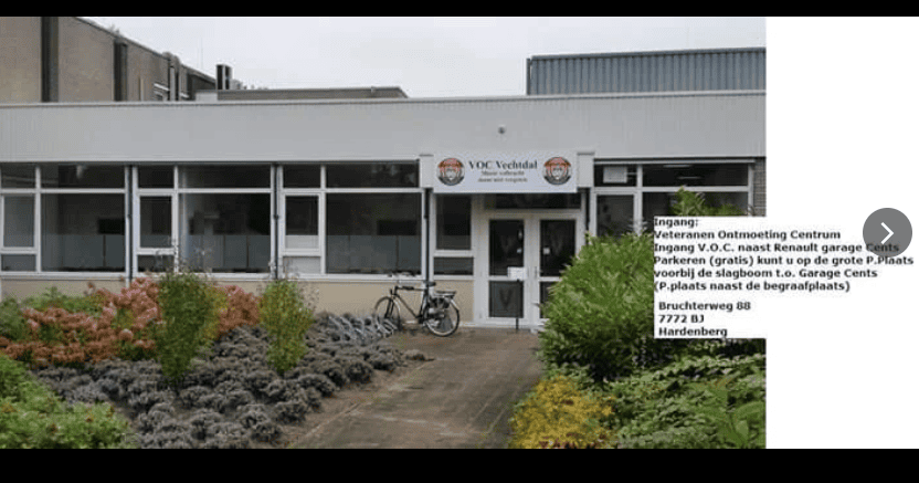 Vooraanzicht van het Veteranen Ontmoeting Centrum Vechtdal in Hardenberg. Voor het gebouw staan struiken en beplanting en bij de ingang staat een fiets. Rechts in beeld staat informatie over de ingang, bereikbaarheid en parkeermogelijkheden.